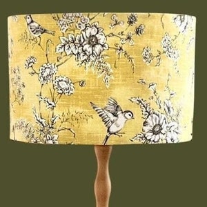 May include: A decorative lamp with a yellow lampshade featuring a floral and bird pattern in black and white. The lamp has a wooden base and a cylindrical lampshade. The background is a solid olive green.