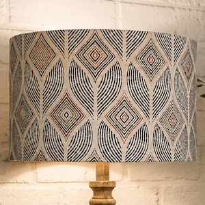 May include: A cylindrical lamp shade featuring a repeating pattern of black, white, and soft coral diamond shapes surrounded by curved, fan-like black lines on a light background, mounted on a textured, light-coloured wooden lamp base.