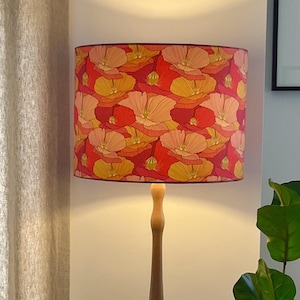 K&ouml;nnte beinhalten: Ein Lampenschirm mit einem floralen Muster in Rot-, Orange- und Gelbt&ouml;nen. Der zylindrische Lampenschirm sitzt auf einem Holzlampenfu&szlig;. Das florale Design zeigt stilisierte Blumen und schafft ein lebendiges, dekoratives Lichtelement.