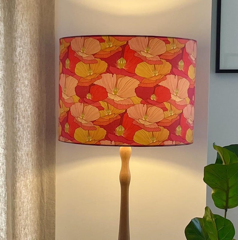 K&ouml;nnte beinhalten: Ein Lampenschirm mit einem floralen Muster in Rot-, Orange- und Gelbt&ouml;nen. Der zylindrische Lampenschirm sitzt auf einem Holzlampenfu&szlig;. Das florale Design zeigt stilisierte Blumen und schafft ein lebendiges, dekoratives Lichtelement.