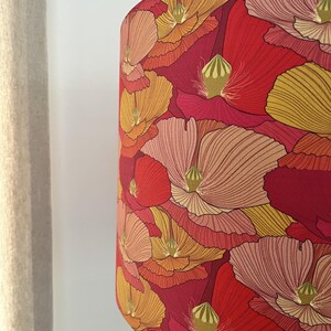 K&ouml;nnte beinhalten: Ein Lampenschirm mit einem auff&auml;lligen Blumenmuster. Das Design zeigt gro&szlig;e, stilisierte Blumen in Rot-, Orange- und Gelbt&ouml;nen vor einem tiefroten Hintergrund. Die Blumen haben detaillierte Bl&uuml;tenblattlinien und gr&uuml;ne Akzente.