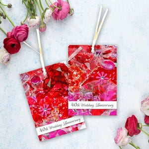 May include: Two red and pink gift tags with a floral design and the text "40th Wedding Anniversary" on a white banner. Each tag has a white ribbon attached for hanging. Flowers are in the background.