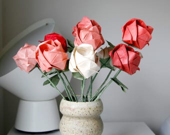 May include: A bouquet of folded paper roses in shades of red, coral, and cream, each featuring intricate geometric petals and green stems. The flowers are arranged in a speckled, light-colored ceramic vase with a rounded, textured body.