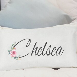 May include: White pillow with the name "Chelsea" written in black cursive script. The name is surrounded by a floral design with pink and green watercolor flowers.