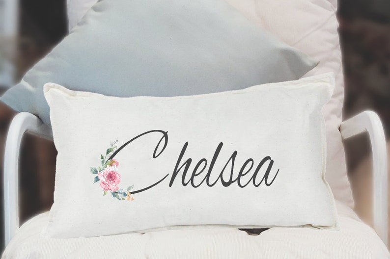 May include: White pillow with the name "Chelsea" written in black cursive script. The name is surrounded by a floral design with pink and green watercolor flowers.