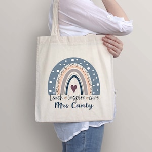 May include: A white canvas tote bag with a rainbow design in shades of blue, pink, and brown. The rainbow has a small heart in the center. The text "teach &hearts; inspire &hearts; care Mrs. Canty" is printed below the rainbow.
