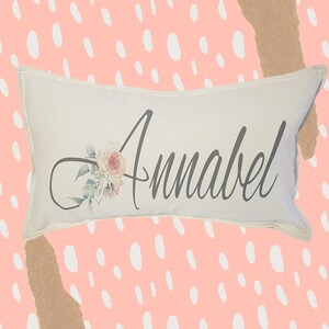 May include: A white pillow with the name "Annabel" printed in gray script with a floral design in pink, white, and green.