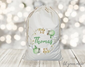 May include: A white canvas gift bag with a green and gold Christmas wreath design. The bag has a drawstring closure and the name "Thomas" is printed in green on the front.