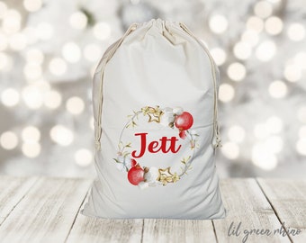 May include: A white canvas Christmas gift bag with a red drawstring. The bag has a watercolor design of a wreath with red ornaments, white cotton, and gold stars. The name "Jett" is written in red inside the wreath.