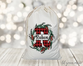 May include: White canvas gift bag with a red and black buffalo plaid letter R and the name Ruben in a banner. The letter R is surrounded by a green wreath with red berries.
