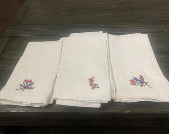 Op de afbeelding: Witte linnen servetten met geborduurde bloemen- en vogelontwerpen in rood, blauw en geel. De servetten zijn gevouwen en gestapeld, waarbij de delicate borduursels op de hoeken te zien zijn. Een klassieke en elegante tafelsetting.