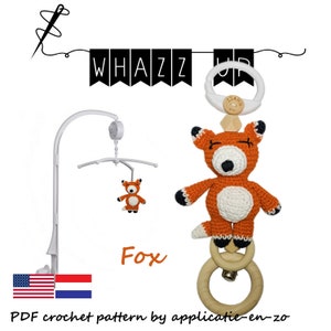 May include: A crochet fox baby mobile with a white frame. The fox is orange with white and black accents. The mobile includes a wooden ring and a bell. The text "Whazz Up" is displayed above the mobile.