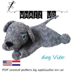 Op de afbeelding: Een gehaakt grijs hondenspeeltje met blauwe ogen en een zwarte neus. De hond ligt met zijn staart omhoog. De tekst "WHAZZ UP" staat boven de hond en "dog Vito" staat eronder. De afbeelding bevat ook de Amerikaanse en Nederlandse vlag.