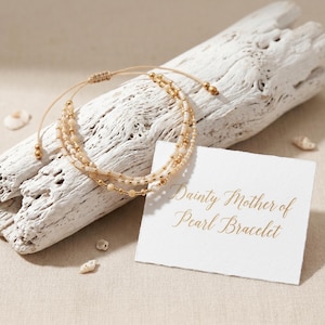 Op de afbeelding: Een delicate, gelaagde armband met kleine, lichtgekleurde kralen en gouden accenten. De armband wordt getoond op een stuk drijfhout, met een kaart met de tekst "Dainty Mother of Pearl Bracelet."