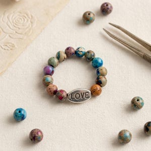 May include: A beaded bracelet with a silver LOVE charm. The bracelet features colorful, speckled beads in shades of blue, green, purple, and orange. Several loose beads and tweezers are scattered around the bracelet on a cream-colored surface.