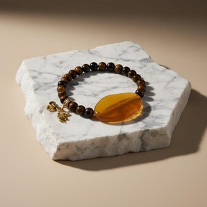 May include: A beaded bracelet featuring brown tiger eye beads and a large amber-colored oval stone. A gold-colored charm with a clover design hangs from the bracelet. The bracelet is displayed on a white marble surface.