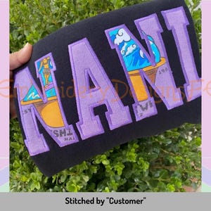 May include: Purple embroidered "NANI" lettering on a black sweatshirt. The letters are outlined in white and feature a cartoon image of a surfer girl on a surfboard.