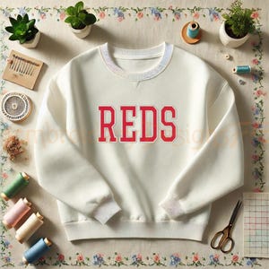 K&ouml;nnte beinhalten: Wei&szlig;er Pullover mit einem roten "REDS"-Logo auf der Vorderseite.