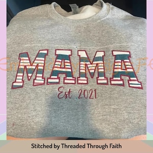 May include: Grey sweatshirt with the word "MAMA" in large, colourful, striped letters. The letters are outlined in maroon. Below the word "MAMA" is the text "Est. 2021" in maroon. The sweatshirt is stitched by "Threaded Through Faith".