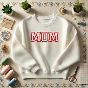 以下が含まれることがあります： クリーム色の長袖スウェットシャツで、赤いブロック体の文字で「MOM」と書かれ、白い水玉模様があしらわれています。リブ編みのクルーネックと袖口が特徴で、裁縫道具に囲まれた平らな場所に置かれています。