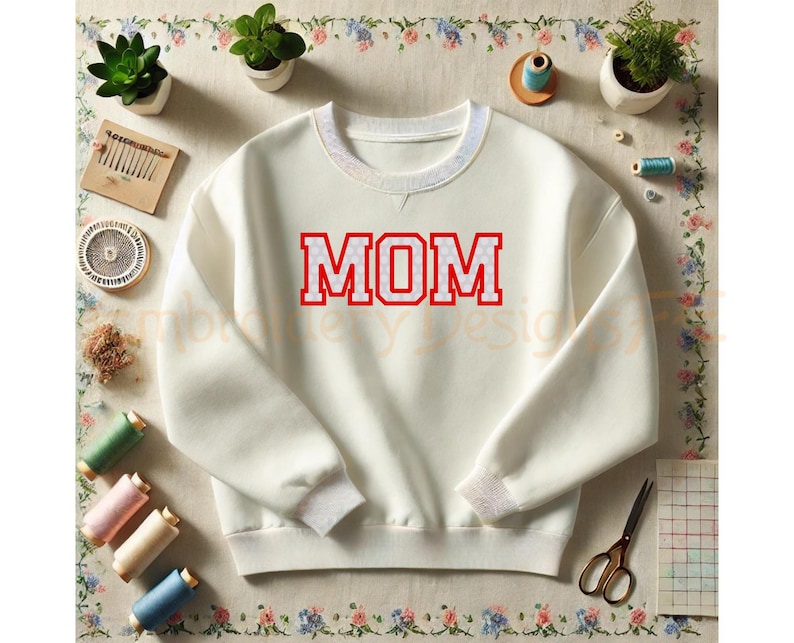 以下が含まれることがあります： クリーム色の長袖スウェットシャツで、赤いブロック体の文字で「MOM」と書かれ、白い水玉模様があしらわれています。リブ編みのクルーネックと袖口が特徴で、裁縫道具に囲まれた平らな場所に置かれています。