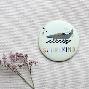 Könnte beinhalten: Ein runder, grün-weißer Knopf mit einem Krokodil-Cartoon, das einen Rucksack trägt und über einen Zebrastreifen geht. Der Text "SCHULKIND" ist in bunten Buchstaben unter dem Krokodil geschrieben.