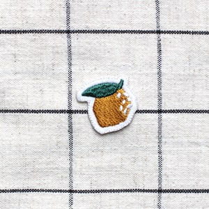 Könnte beinhalten: Ein kleiner gestickter Aufnäher mit einer Orange und einem grünen Blatt auf einem weißen und schwarzen karierten Stoffhintergrund.