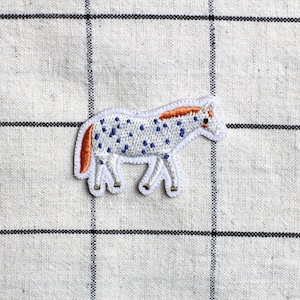 Könnte beinhalten: Weißer, gestickter Bügeleisen-Aufnäher mit einem gepunkteten Pferd mit orangefarbener Mähne und Schweif. Das Pferd blickt nach rechts und hat ein blaues und weißes Punkte-Muster.