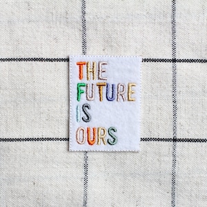 Può includere: Un toppa di tessuto bianco con il testo "THE FUTURE IS OURS" ricamato in filo colorato.