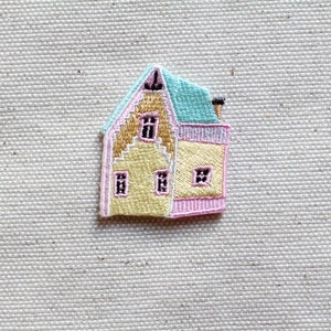 Può includere: Una piccola toppa ricamata di una casa gialla con accenti rosa e blu. La casa ha tre finestre e un camino.
