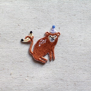 Op de afbeelding: Geborduurde patch met een bruine aap met een paarse feesthoed met een blauwe pompon. De aap houdt een banaan vast. De patch is gemaakt met gedetailleerde steken en heeft een witte rand. Op een beige stof.