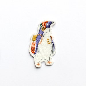May include: Embroidered iron-on patch featuring a penguin wearing a purple and orange backpack. The penguin is white with a black beak and black eyes. The patch is approximately 5 cm tall.