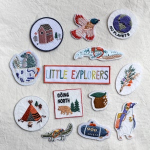 May include: A set of 11 embroidered iron-on patches featuring various designs, including a house, a bird, a compass, a tree, a tent, a submarine, a penguin, and the text "Little Explorers".