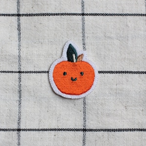 May include: A smiling orange embroidered pumpkin with a green stem and brown stalk on a white and black checkered background.