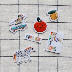 May include: Five embroidered iron-on patches with various designs. The patches include a penguin on a skateboard, a red apple with a smiling face, a lemon, a zebra, and a white patch with the text "The Future Is Ours" in rainbow colours.