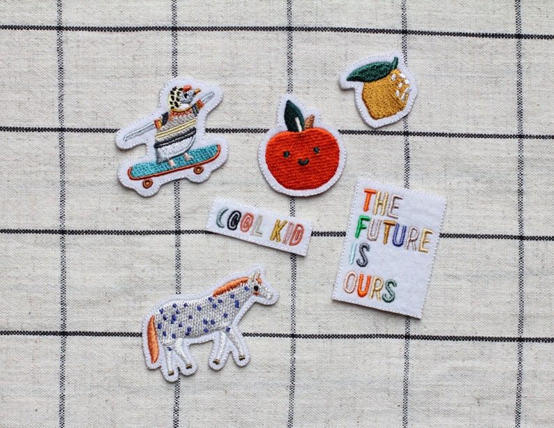 May include: Five embroidered iron-on patches with various designs. The patches include a penguin on a skateboard, a red apple with a smiling face, a lemon, a zebra, and a white patch with the text "The Future Is Ours" in rainbow colours.