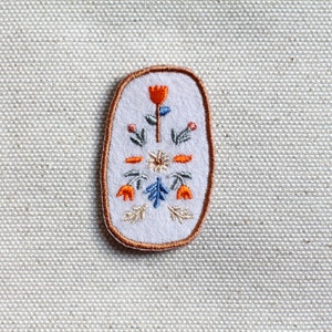 Könnte beinhalten: Ein weißer ovaler gestickter Aufnäher mit einem Blumenmuster. Das Design zeigt eine zentrale orangefarbene Blume mit zwei kleineren orangefarbenen Blumen auf beiden Seiten. Die Blumen sind von grünen Blättern und blauen Akzenten umgeben.
