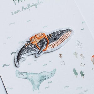 Könnte beinhalten: Eine weiße Karte mit dem Text "Papa zum Aufbrügeln" und einem kleinen gestickten Aufnäher einer Wal mit einer orangefarbenen Schwimmweste. Der Wal schwimmt in einem blauen Meer mit weißem Hintergrund.
