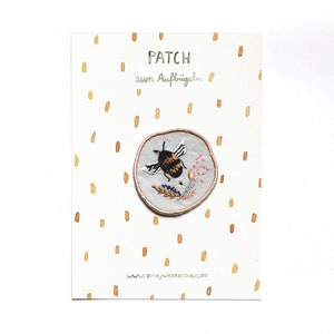 May include: A white fabric patch with a round embroidered design of a bumblebee flying over flowers. The text "PATCH zum Aufb&uuml;geln" is printed on the card.