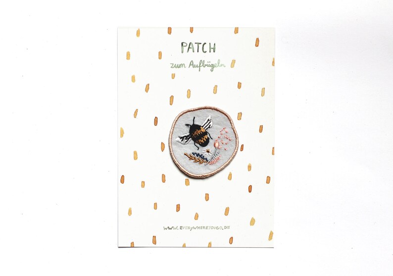 May include: A white fabric patch with a round embroidered design of a bumblebee flying over flowers. The text "PATCH zum Aufb&uuml;geln" is printed on the card.