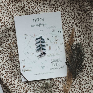 Könnte beinhalten: Eine weiße Karte mit einer handgezeichneten Illustration einer Waldszene. Die Karte sagt "PATCH zum Aufbügeln" und "SOUTA, IVER und der Wind". Auf der Karte befindet sich ein grüner gestickter Aufnäher mit einer Kiefer.