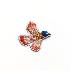 Op de afbeelding: Een klein geborduurd pleister met een bruine en oranje vogel met blauwe accenten, vliegend met zijn vleugels uitgespreid.