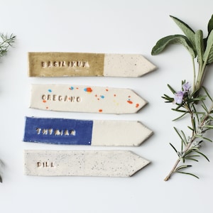 Op de afbeelding: Vier keramische plantenlabels met pijlvormige uiteinden. De labels zijn geëtiketteerd met "Basilicum", "Oregano", "Tijm" en "Dille".