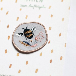 May include: A round grey embroidered patch featuring a black and yellow bee with white wings and floral details. The patch has a brown outline and says "Patch zum Aufb&uuml;geln" in black text.
