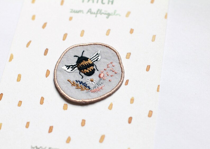 May include: A round grey embroidered patch featuring a black and yellow bee with white wings and floral details. The patch has a brown outline and says "Patch zum Aufb&uuml;geln" in black text.