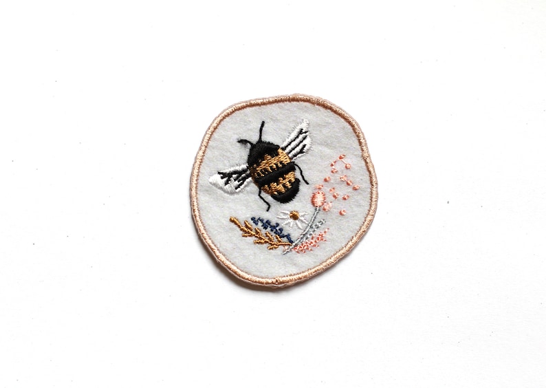 May include: A round embroidered patch featuring a black and gold bee surrounded by pink and white flowers. The patch has a light brown border.