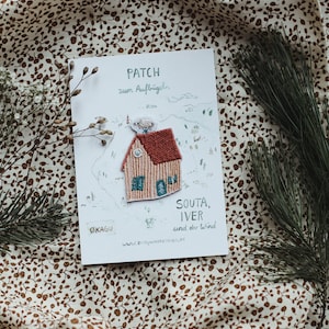 Può includere: Un biglietto bianco con il testo "PATCH zum Aufbügeln" e "SOUDA, IVER und der Wind" con una toppa ricamata di una casa rossa e gialla in cima. Il biglietto è su un tessuto bianco con un motivo floreale marrone.