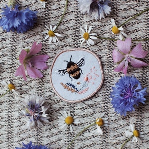 May include: A white embroidered patch featuring a bee and flowers, set against a beige fabric background. The patch is surrounded by a variety of flowers, including daisies, cornflowers and pink flowers.