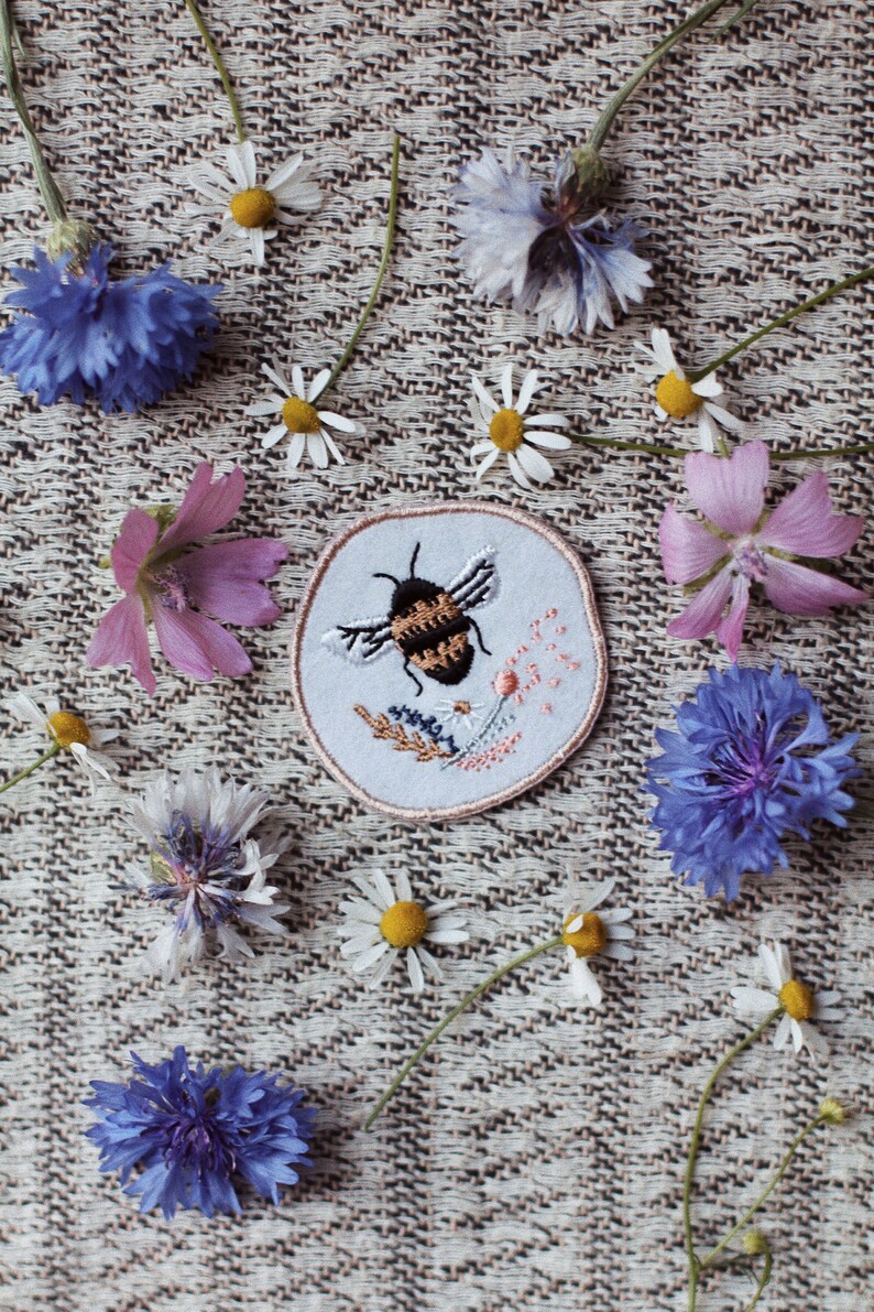 May include: A white embroidered patch featuring a bee and flowers, set against a beige fabric background. The patch is surrounded by a variety of flowers, including daisies, cornflowers and pink flowers.