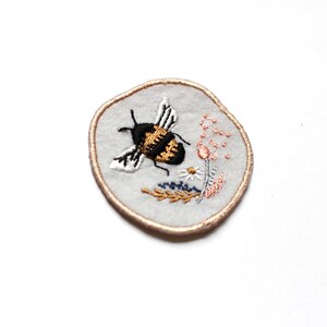 May include: A round embroidered patch with a white background and a brown outline. The patch features a black and yellow bee with white wings flying over a pink flower and green leaves.